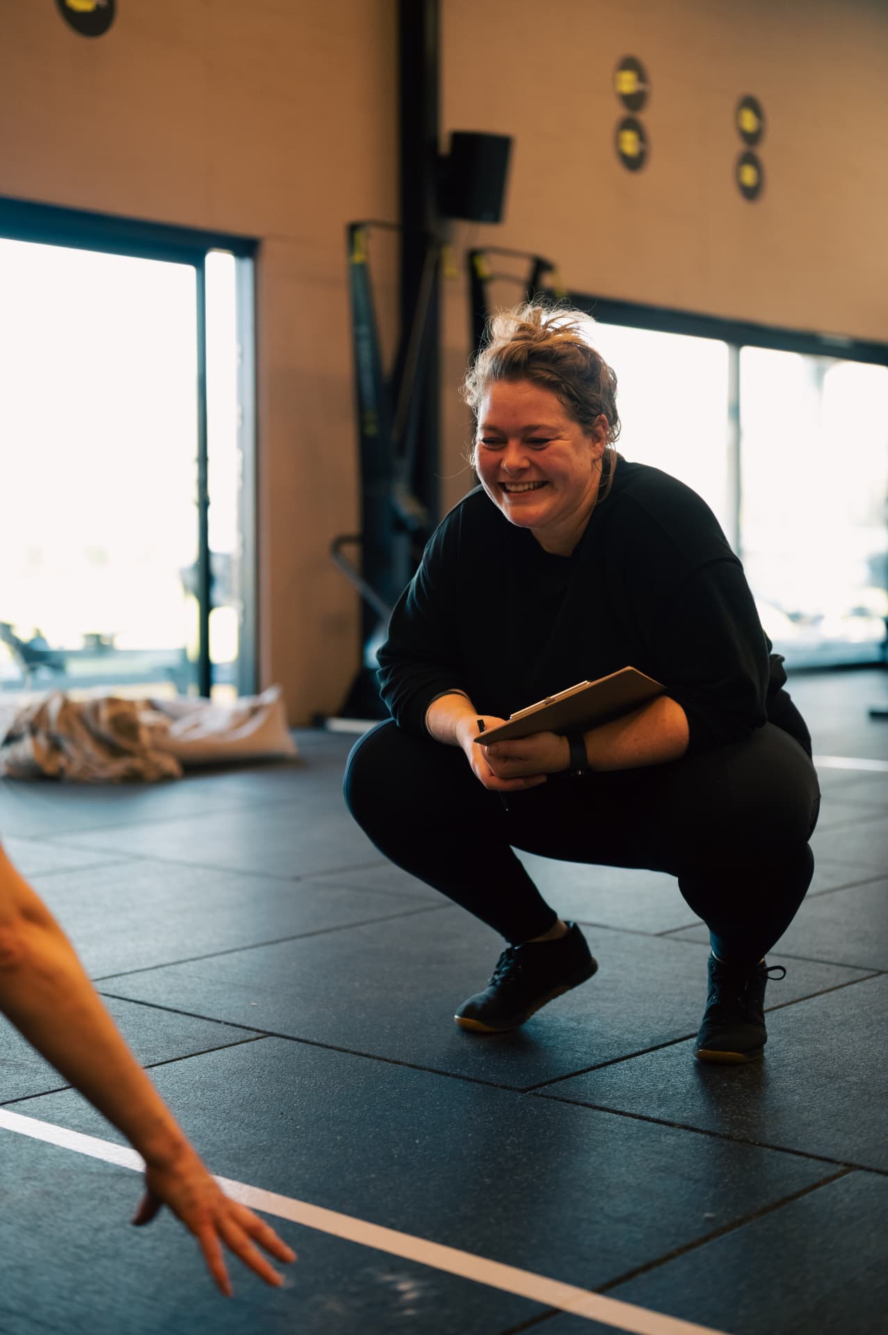 A smiling coach crouching with a clipboard, encouraging a member mid-workout