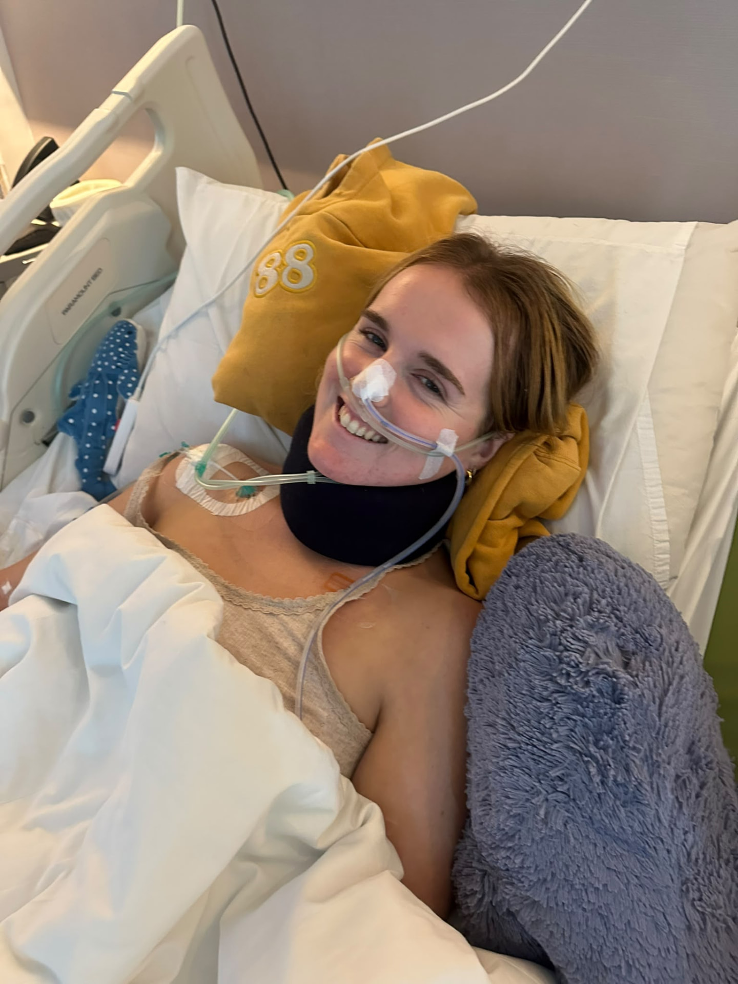 Georgia smiling in a hospital bed beside a soft toy, wearing a neck brace and nasal tube