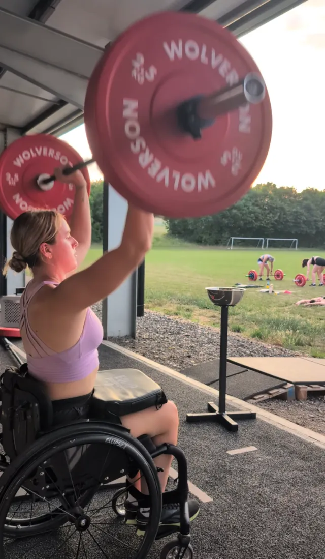 Georgia lifting a barbell overhead from her wheelchair at the gym