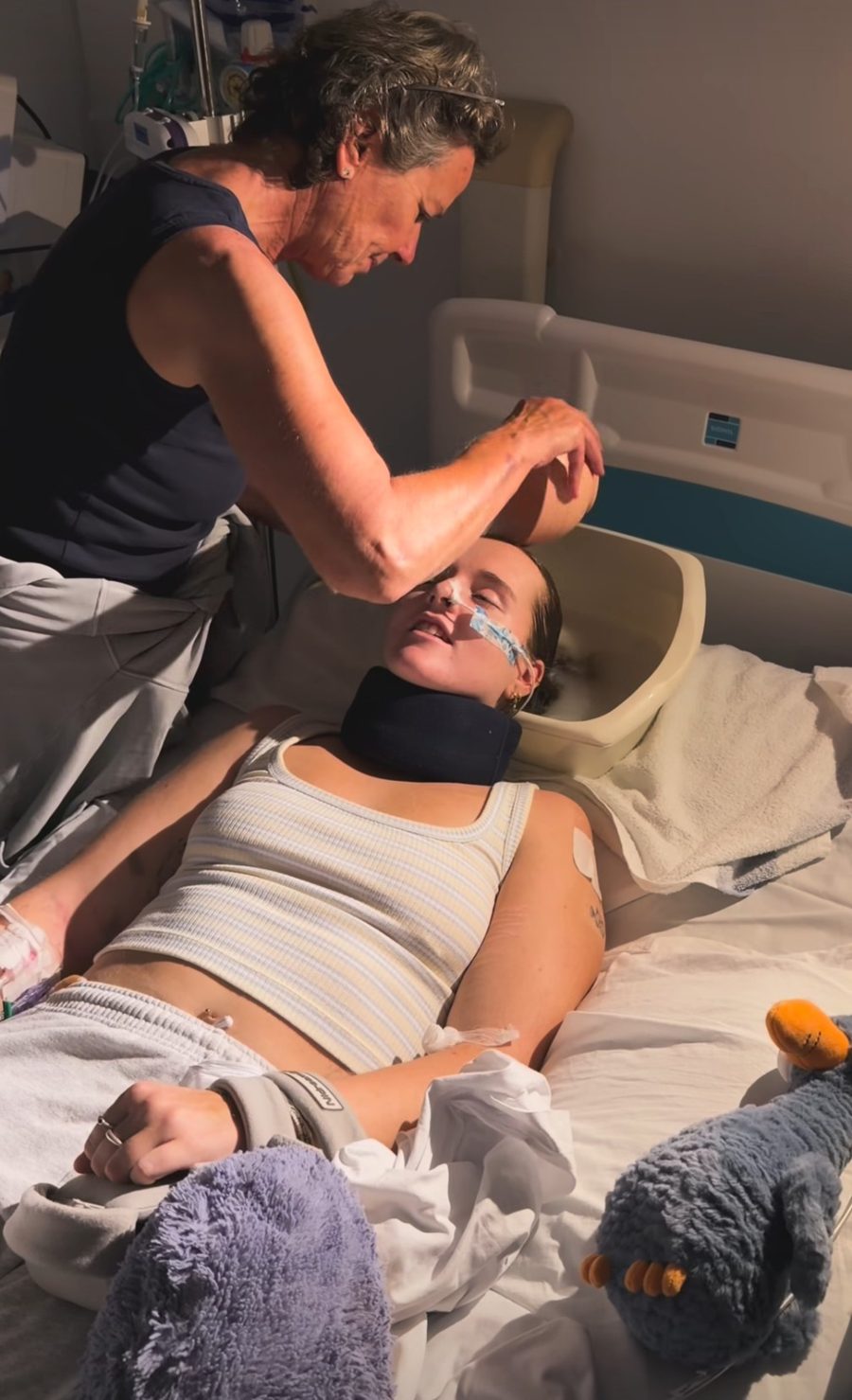 Georgia lying in a hospital bed wearing a neck brace, with someone gently washing her hair.